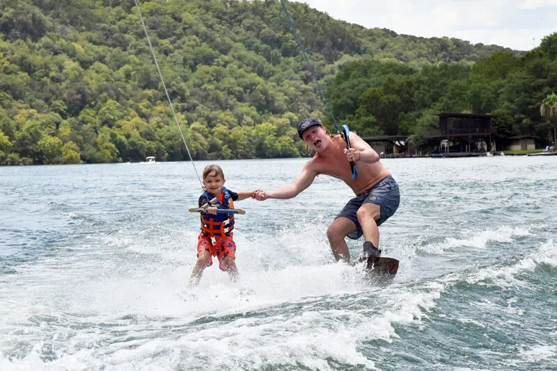 Adult teaching a child to wakeboard on a lake with forested hills in the background
